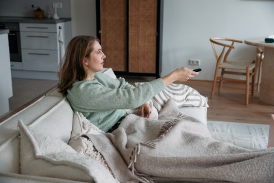 frau liegt auf couch mit decke und fernbedienung in der hand