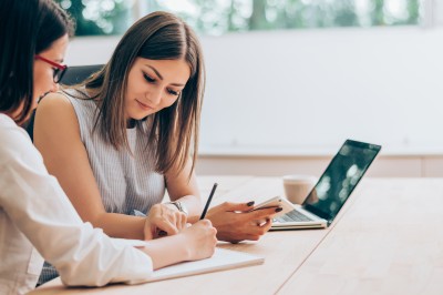zwei frauen sitzen am schreibtisch und notieren sich informationen