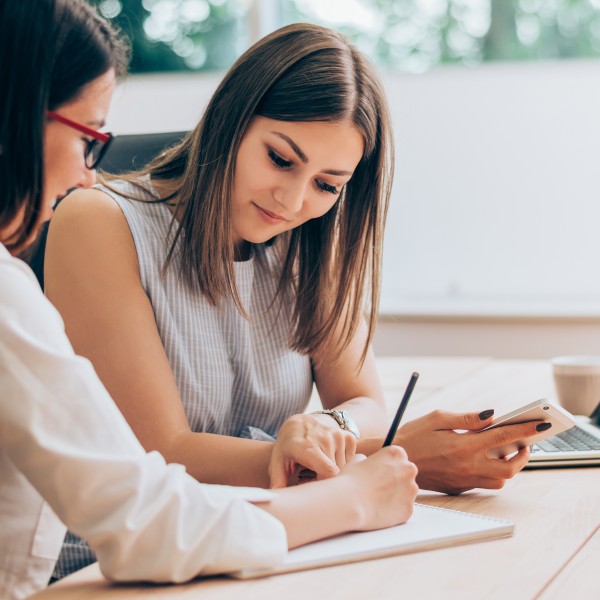 zwei frauen sitzen am schreibtisch und notieren sich informationen